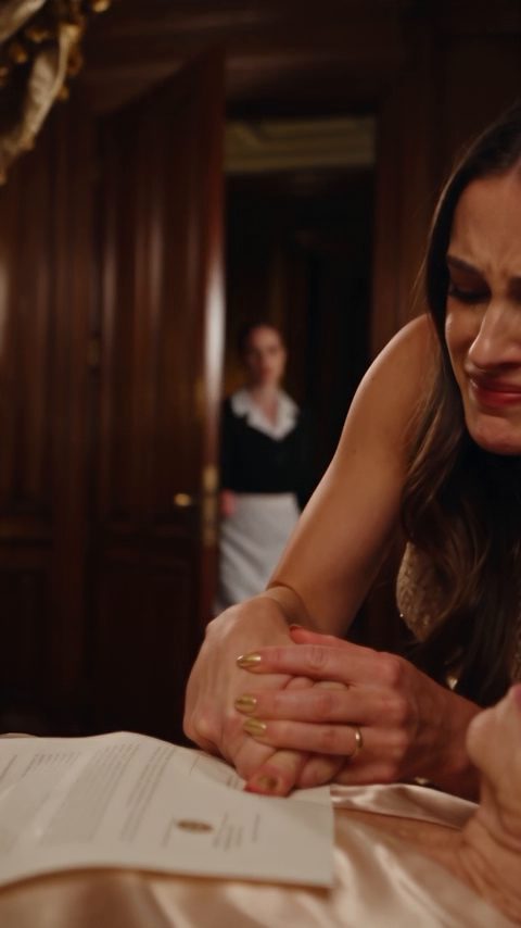 A distressed woman in a gold dress looks toward a maid standing in the dark mansion doorway.